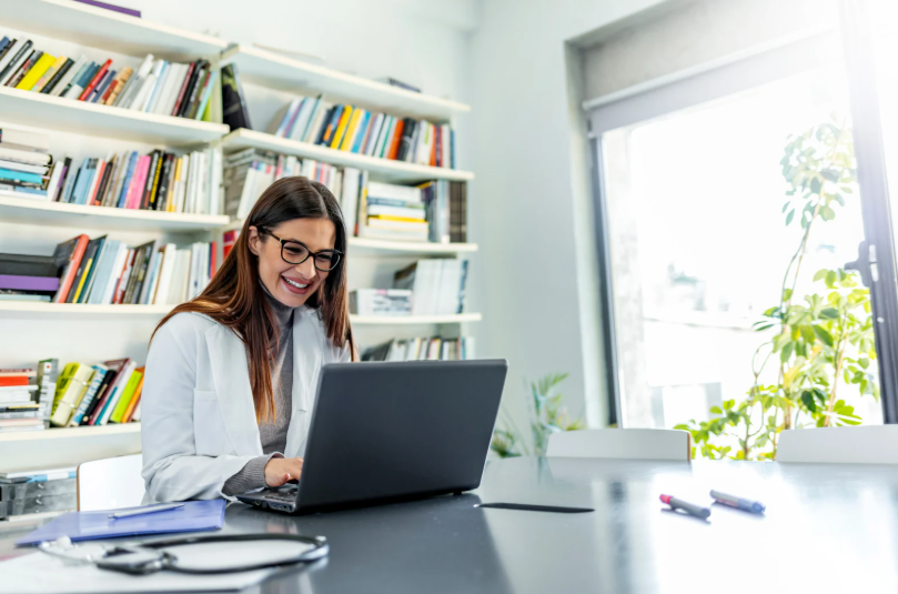 A woman in a white lab coat, sitting at a desk with a laptop, smiling while working. She is surrounded by bookshelves and has glasses on, creating a professional and focused environment.
